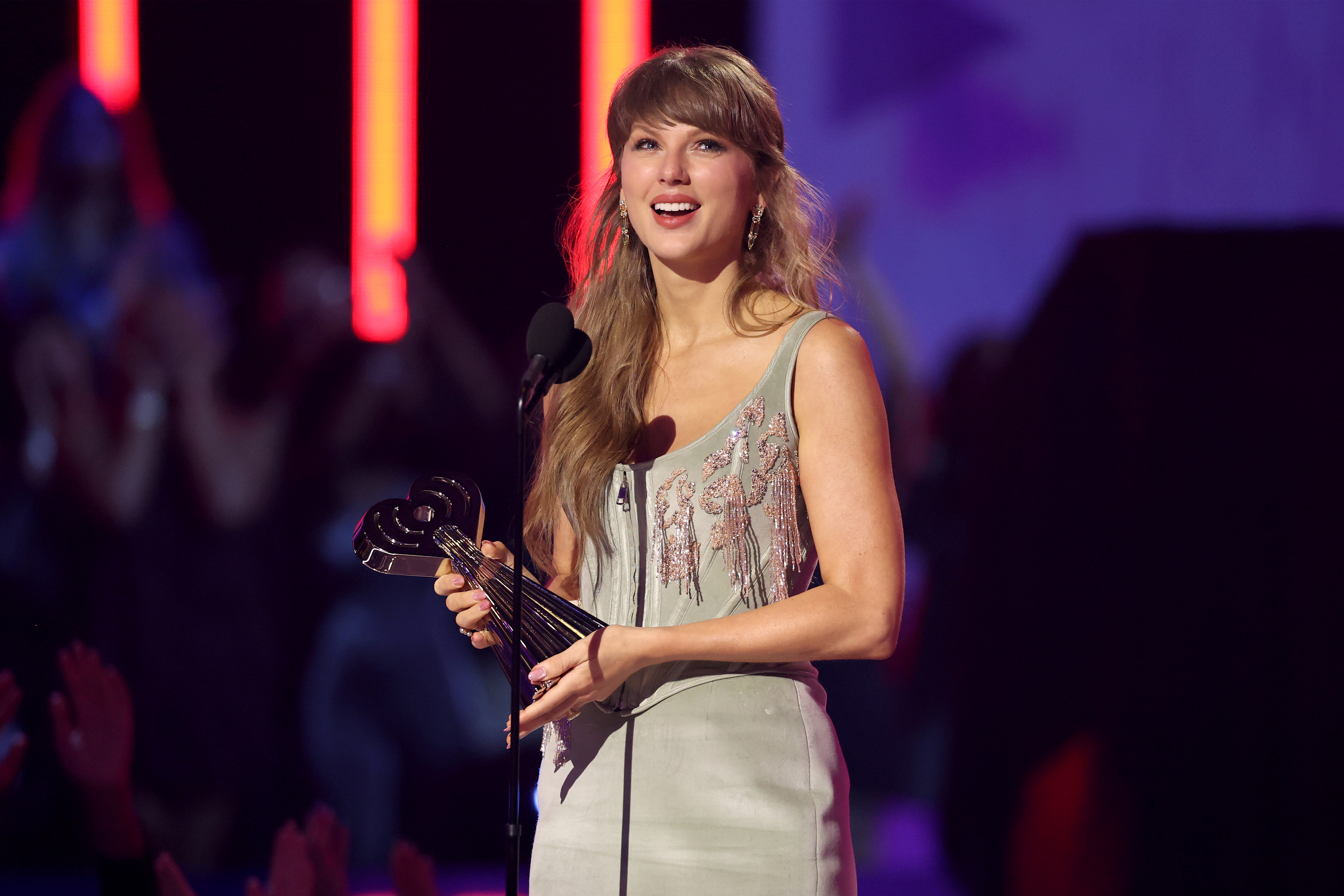 Taylor Swift on stage performing with a guitar, a pink bodysuit, and silver boots, lit by stage lights.